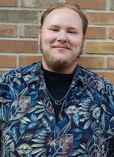 Headshot of Ulysses Swanson, September 2024. Photo of a man with pink-white skin, red short hair and a beard, and a black and maroon floral button up in front of a brick wall.