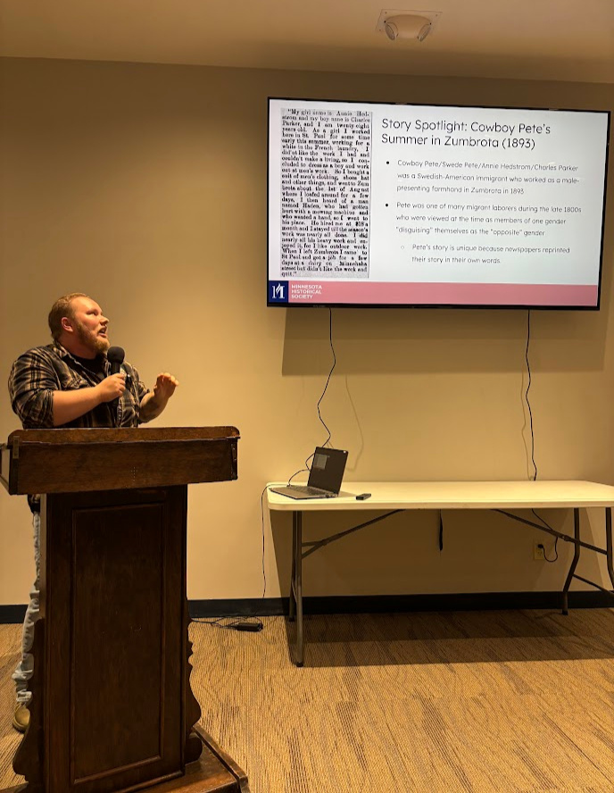 Ulysses Swanson, representing MNHS, at the Goodhue County Historical Society, Red Wing, MN. June 13, 2025. Photo of a white man in a brown plaid shirt standing at a podium and holding a microphone. He is staring at a screen that shows an image of an old-timey newspaper and text.