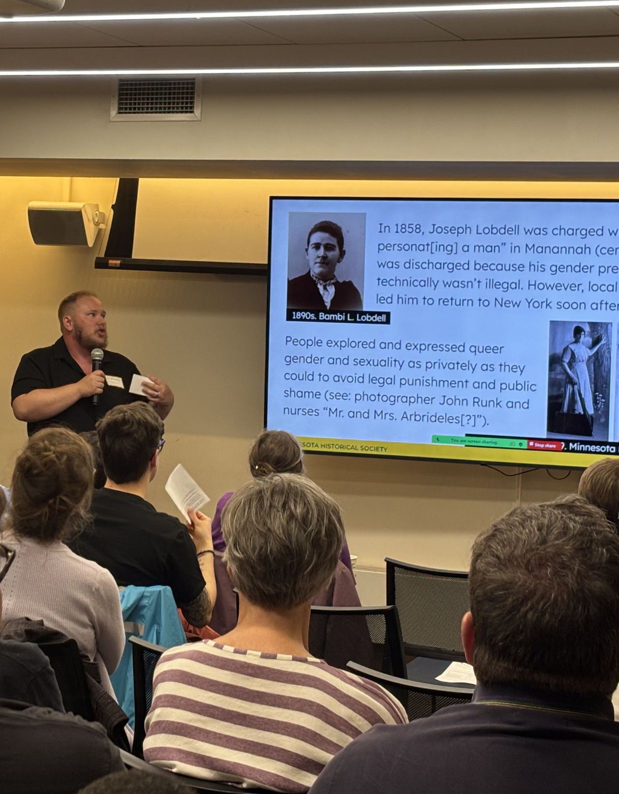 Photo of a white man in a black button up shirt standing and holding a microphone. He is staring at a screen that shows two historic photographs of gender nonconforming people.